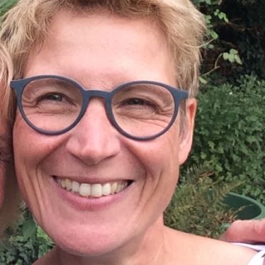 Passfoto, Frau mit kurzen blond/braunen Haaren, mit blauer Brille, lachend, grüne Sträucher im Hintergrund